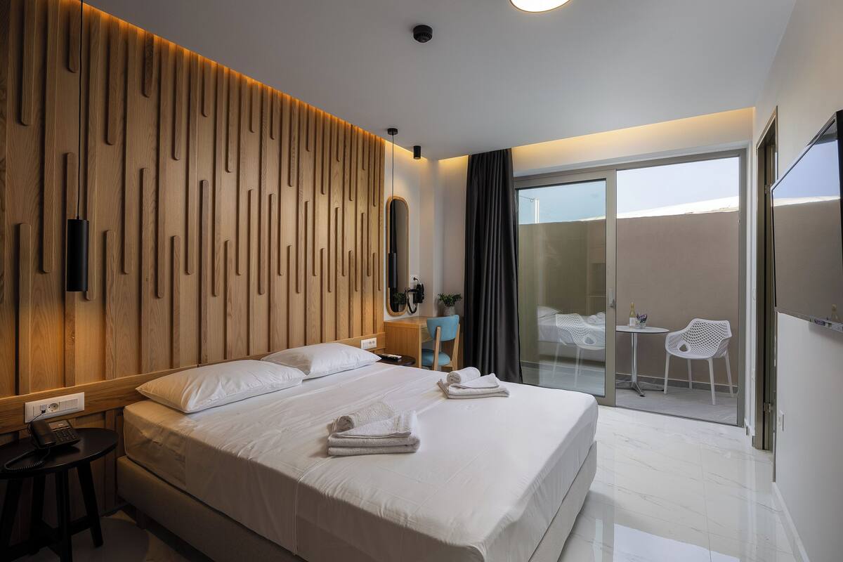 A modern hotel room with a large bed, a desk with a phone, a chair, a small table with a plant, and a balcony with a chair and table.