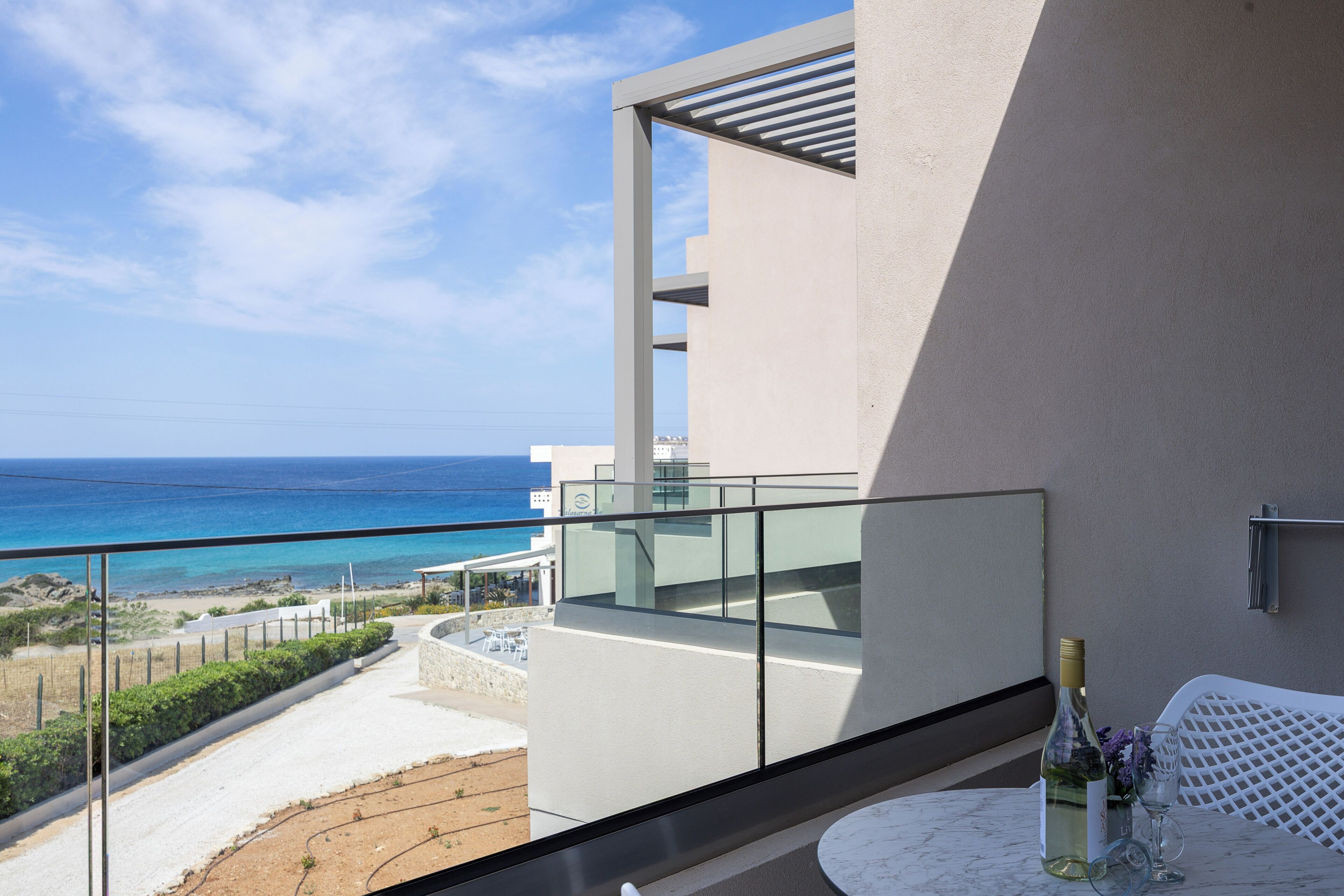 A balcony with a clear view of the ocean, a white table with a bottle and glass, and a modern building with a glass railing.