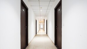 Um corredor comprido e estreito, com portas de madeira escura em ambos os lados, paredes brancas e chão de azulejos.