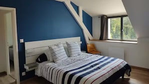 Un dormitorio con techo inclinado, cama con ropa de cama a rayas, una silla y una ventana con vista a árboles.