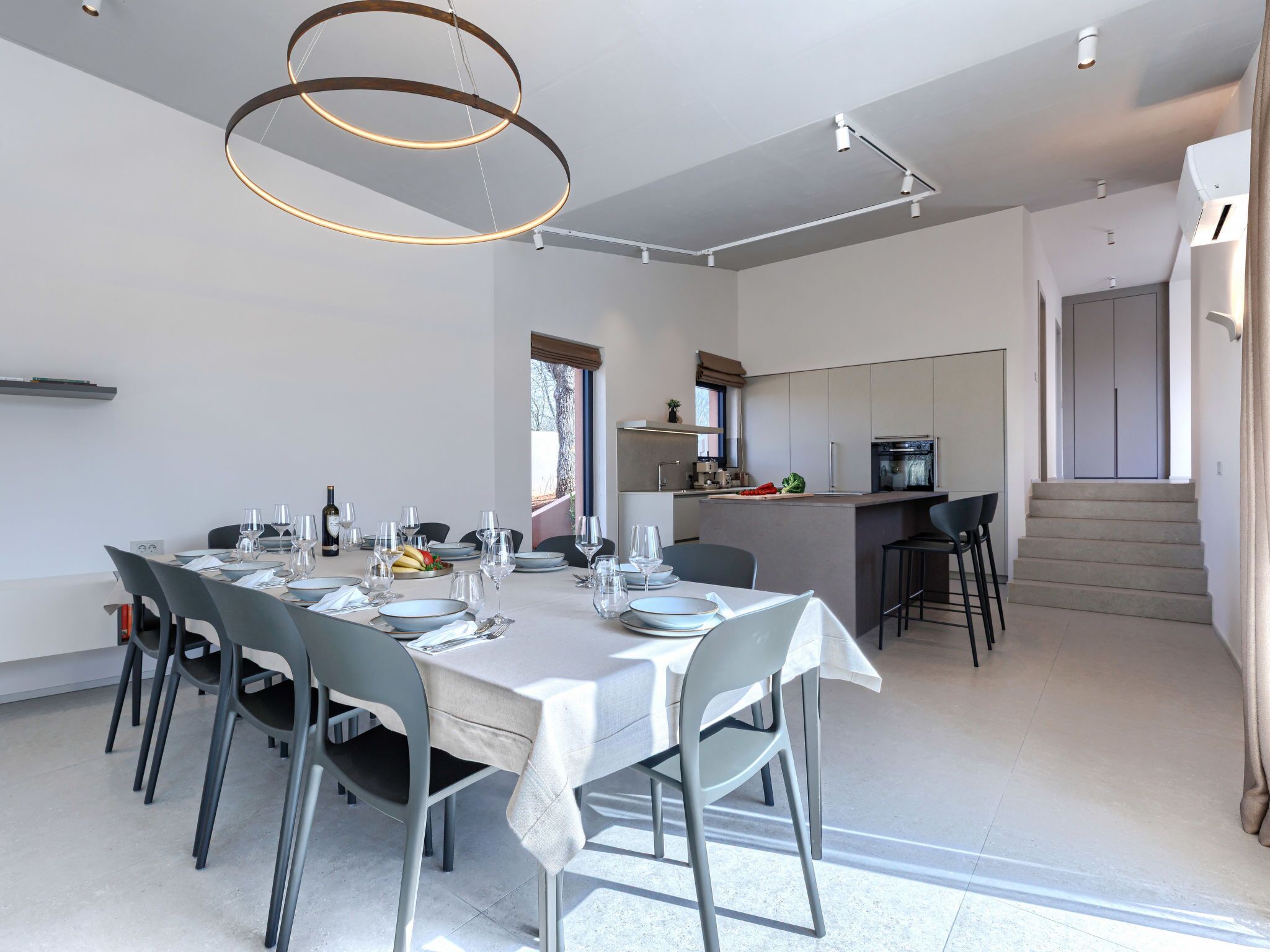 Un espace de repas moderne avec une longue table dressée pour six personnes, une cuisine en arrière-plan et un escalier menant à un niveau supérieur.