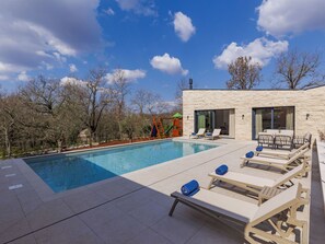 Piscina con sillas de descanso y tobogán, rodeada de árboles y un edificio moderno.