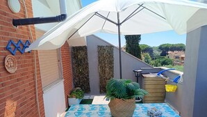 Eine kleine Terrasse mit einem Tisch, bedeckt mit einer blau-blumigen Tischdecke, einem weißen Sonnenschirm und einem Blumentopf.