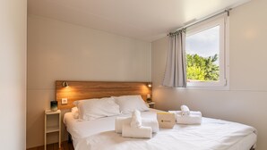Ein Hotelzimmer mit einem hölzernen Kopfteil, einem Bett mit weißen Leinen, einem Nachttisch mit einer Lampe und einem Fenster mit Blick auf Grünflächen.