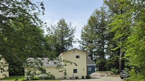 A two-story house with a driveway leading to a gated entrance, surrounded by lush greenery and trees.