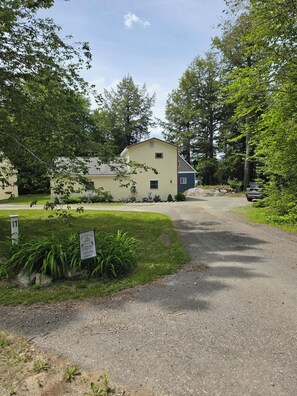 Une maison à deux étages, avec une allée menant à une entrée fermée par un portail, entourée d’une végétation luxuriante et d’arbres.