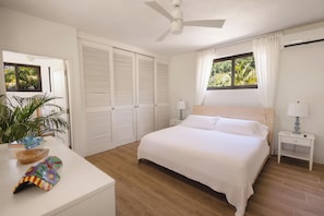 Un dormitorio con cama, mesitas de noche, ventilador de techo, ventana con cortinas y puerta con vista a follaje.