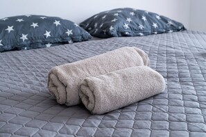 Una cama con una colcha gris acolchada, dos almohadas con motivos de estrellas y dos toallas enrolladas.