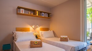 A hotel room with two beds, a shelf with books and decorative items, a small table with a vase, and a towel with 'Club del Sole' branding. - Tenuta Primero Grado Family Collection (Grado)