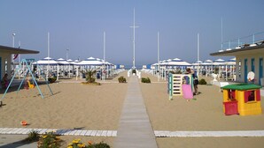 A beachside promenade with rows of beach umbrellas, a children's play area, and beach chairs. - Tenuta Primero Grado Family Collection (Grado)