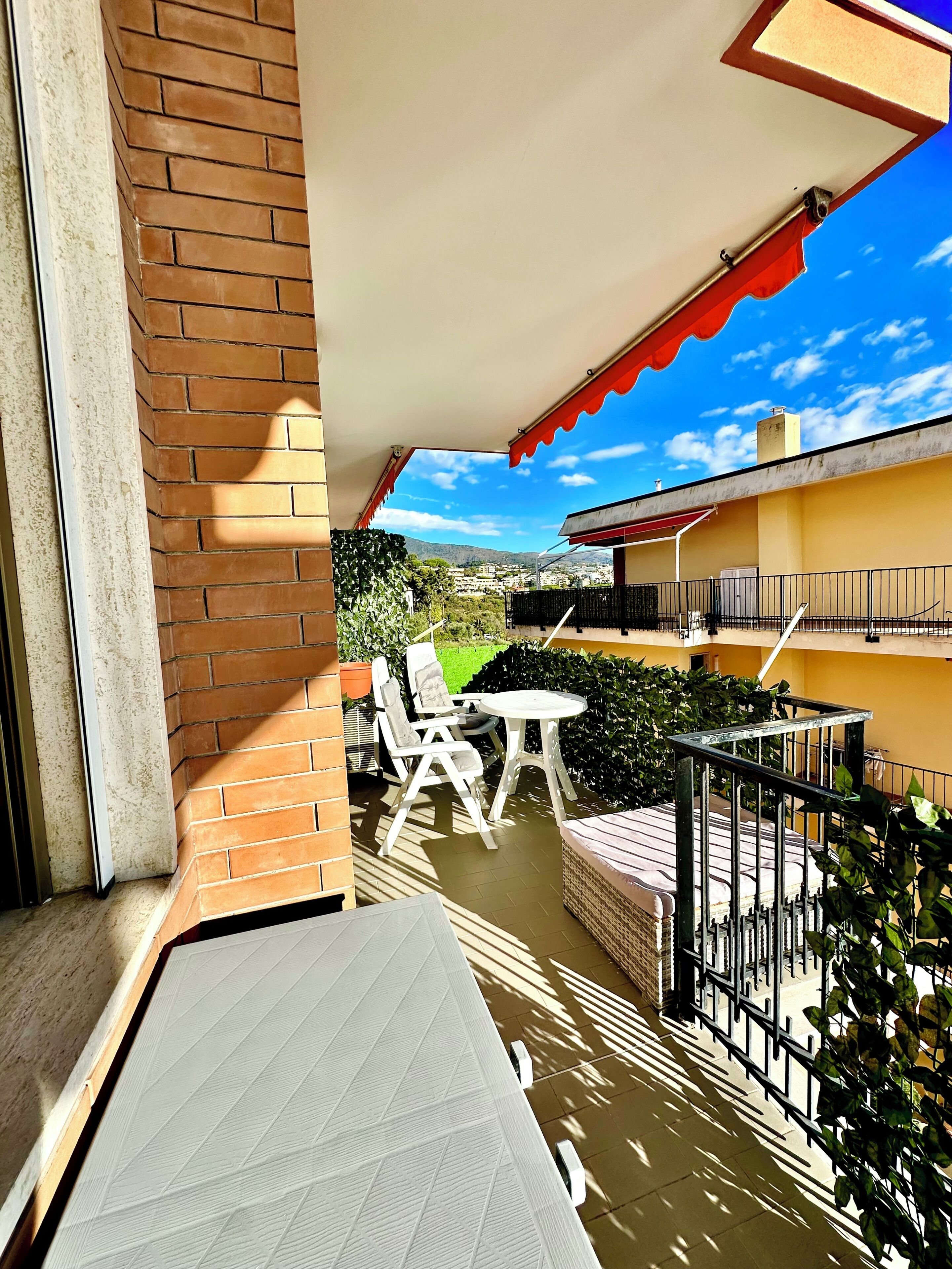 A balcony with a white table and chairs, a red awning, and a view of a yellow building and greenery.