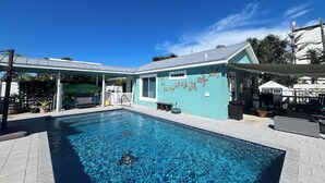 Una piscina cristallina con una zona patio coperta, una casa con facciata color acquamarina e una recinzione.