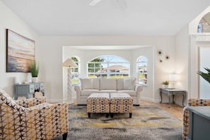 A living room with a sofa, ottoman, and patterned chairs, a window with a view, and a ceiling fan. - Feel @ Home Ft Myers - 3bd - 2bh - POOL - Sleeps 8 (Fort Myers)