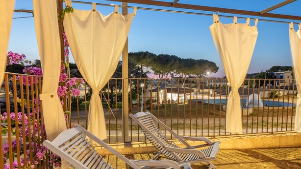 A covered outdoor seating area with two lounge chairs and white curtains, overlooking a landscaped garden and a pool. - Le Terrazze Del Sud - Salve (Salve)