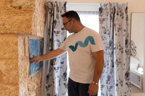 A man interacting with a wall-mounted device, possibly a thermostat, in a room with floral curtains and a white wall.