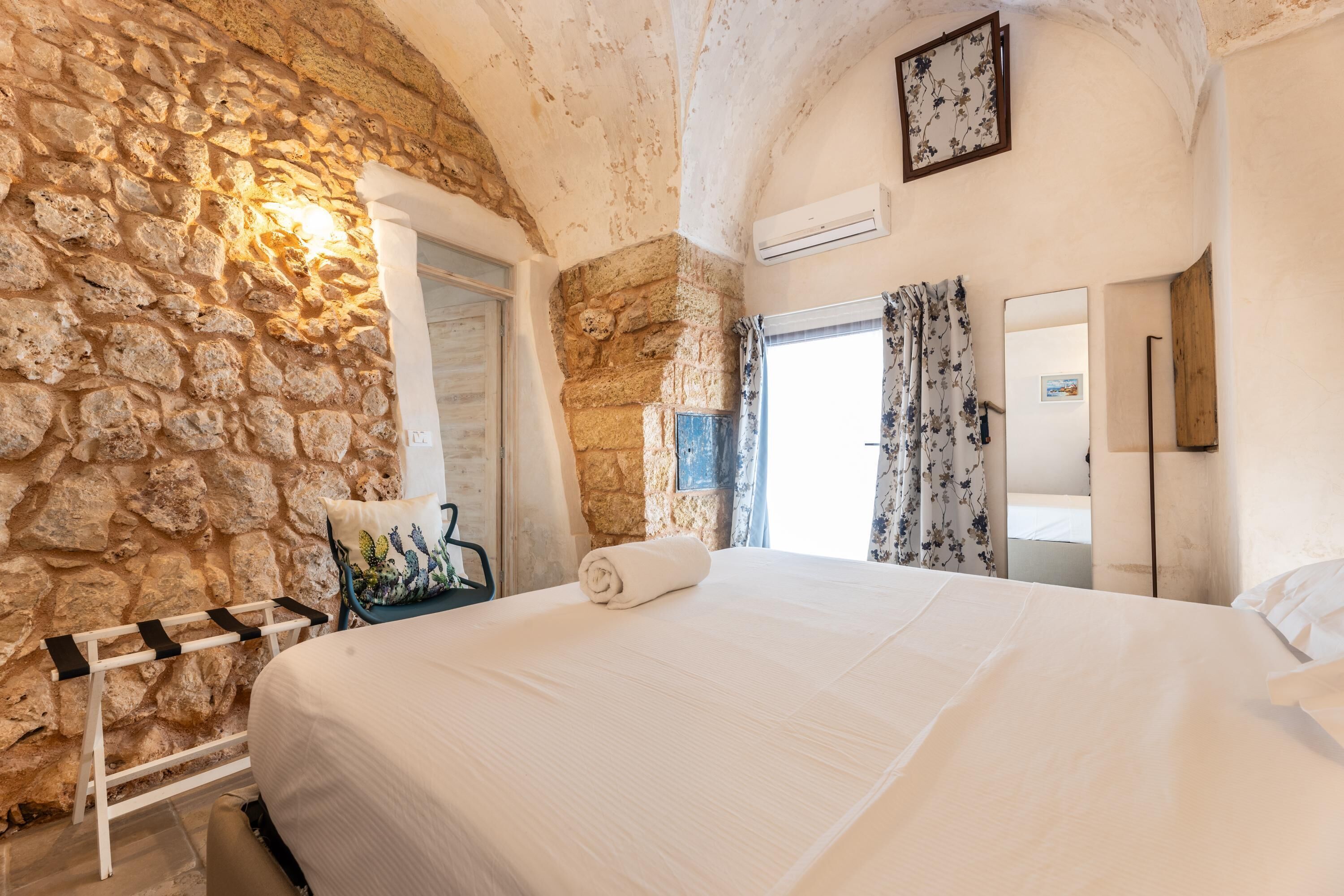 A bedroom with a stone wall, a bed with a rolled towel, a chair with a cactus pillow, and a door with a picture.