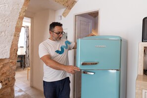 A man opening a vintage teal refrigerator in a room with a stone wall and a door leading to another room. - Tenuta Terra E Sale - Salve (Salve)