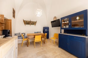 A compact kitchen with a dining area, a blue cabinet with glass doors, a vintage coffee machine, and a wall-mounted TV. - Tenuta Terra E Sale - Salve (Salve)