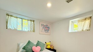 Un dormitorio con cama, almohadas, un cuadro en la pared y dos ventanas con cortinas.