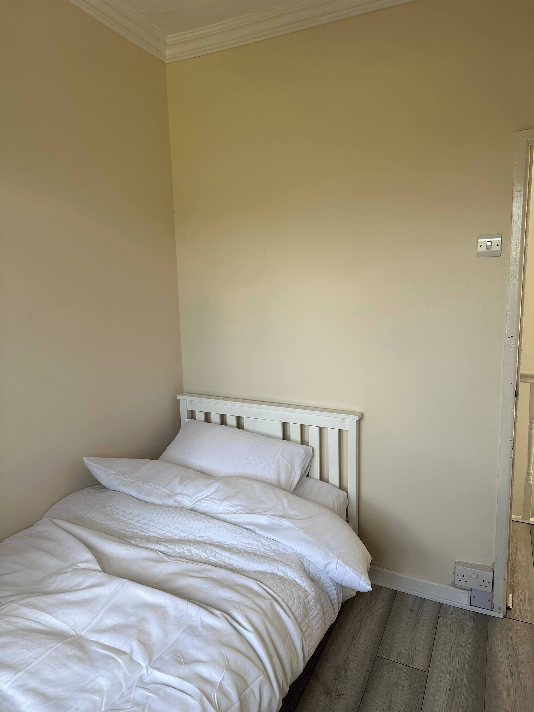 A bedroom with a bed, white bedding, a wooden headboard, a light switch, and a power outlet on the wall.