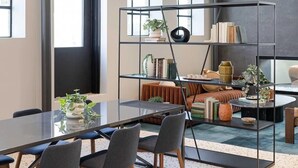 A modern living room with a glass dining table, grey chairs, a sofa, and a bookshelf. - Sleepover | Elite Studio + Gym/Sauna - Omaha (Omaha)