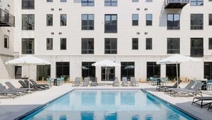 Une piscine entourée de chaises longues et de parasols, le tout bordé d’un bâtiment doté de nombreuses fenêtres et de balcons.