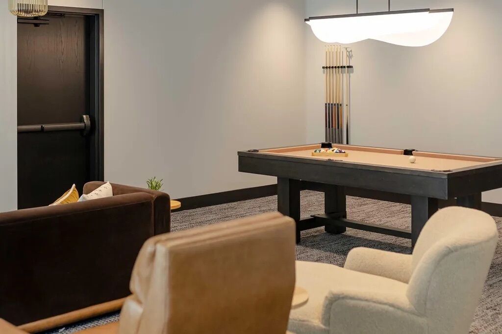 Ein Billardtisch mit einer darüber hängenden Leuchte, eine braune Couch und ein weißer Sessel in einem Raum mit grauem Teppich.