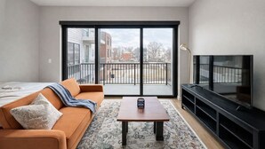 Een moderne woonkamer met een bank, een salontafel, een televisie en een balkon met uitzicht op gebouwen.