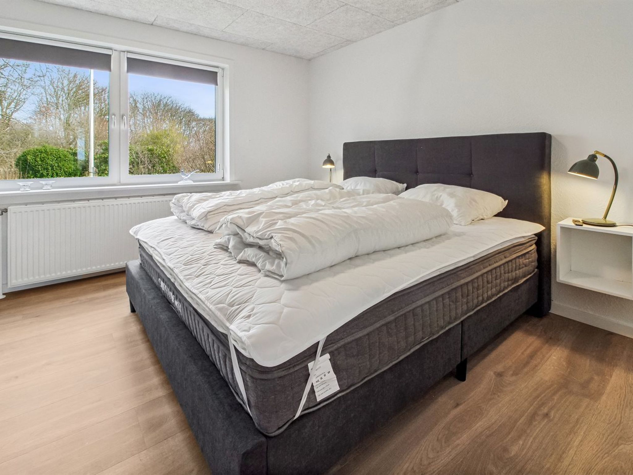 Et soveværelse med en stor seng, et natbord med lampe, et vindue med udsigt til træer og en hvid radiator.