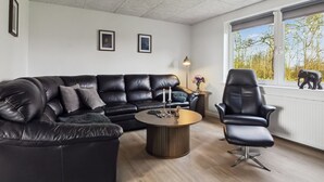 A modern living room with a black leather sofa, a round coffee table, and a black leather armchair. There are two framed pictures on the wall, a floor lamp, and a window with a view of trees and an elephant statue. - "Milan" - 20km from the sea by Interhome (Bredebro)