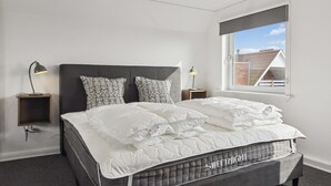 A neatly made bed with a quilted mattress, two patterned pillows, and a headboard. There are bedside tables with lamps, a window with a view of buildings, and a carpeted floor. - "Milan" - 20km from the sea by Interhome (Bredebro)