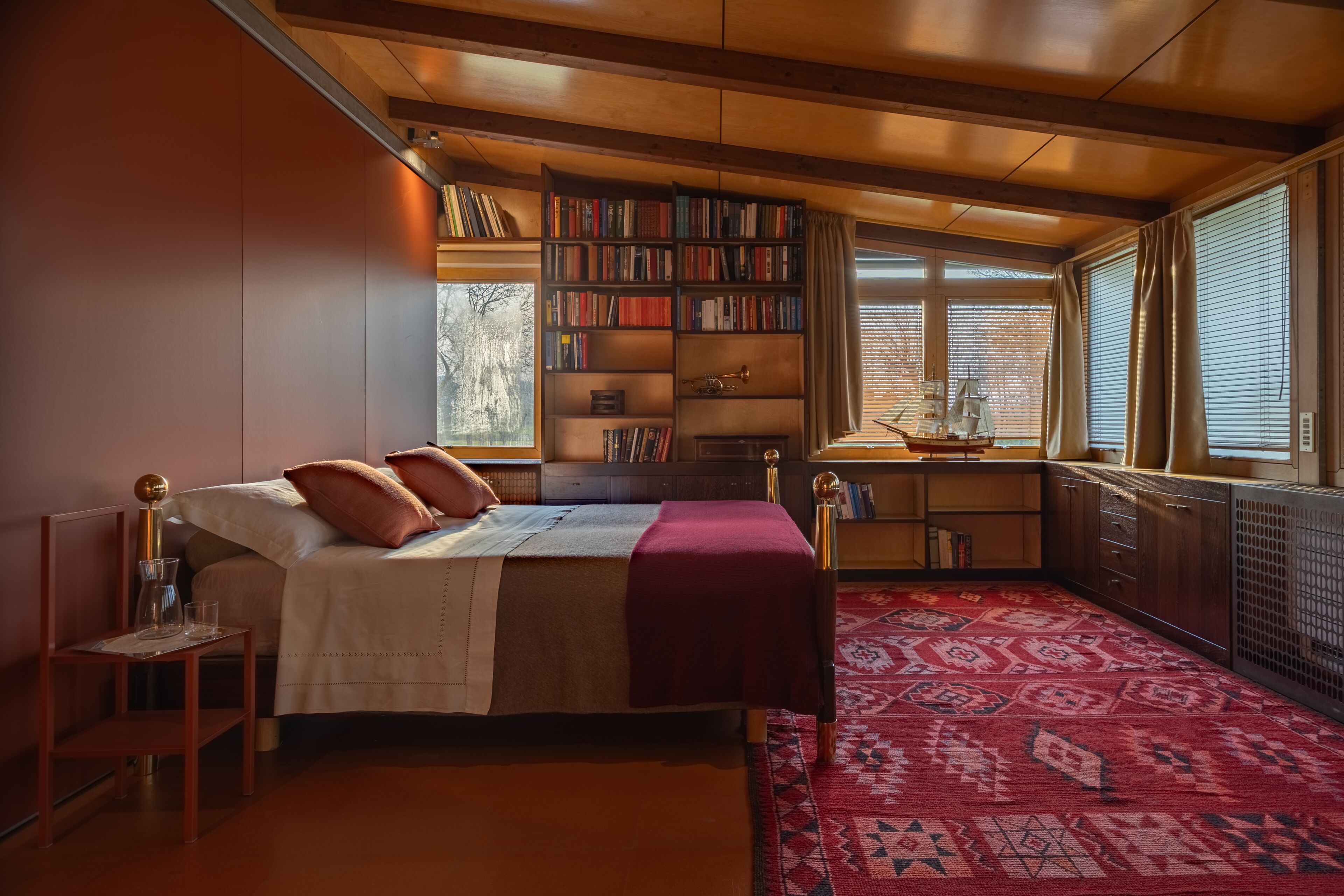 A bedroom with a large bed, a bookshelf, a desk with a ship model, and a patterned rug.