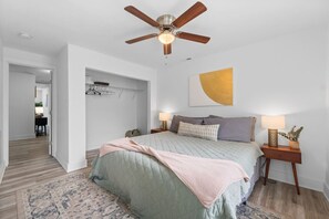 Ein Schlafzimmer mit einem Bett, Nachttischen, einem Deckenventilator, einem Wandbild und einem Flur, der in einen anderen Raum führt.