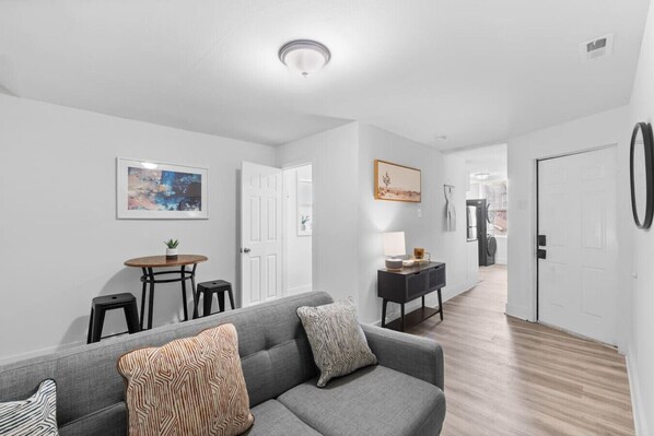 A modern living room with a gray sofa, a small dining table, and a hallway leading to a kitchen.