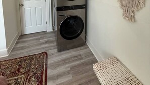 Une buanderie équipée de deux machines à laver et de deux sèche-linge superposés, une porte blanche, un tapis à motifs et une décoration murale en macramé.