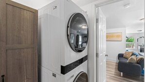 Un lavadero pequeño con lavadora y secadora de carga frontal, una puerta de madera y vistas a una sala de estar con sofá y un cuadro.