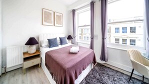 Quarto com cama, mesas de cabeceira, uma cadeira, janela com cortinas e vista para edifícios.