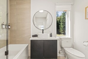 A bathroom with a bathtub, a sink with a mirror, a toilet, and a window with a view of trees.