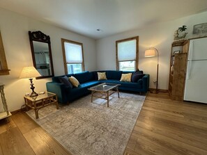 Un salon avec un canapé bleu, une table basse en verre, un tapis à motifs, un parquet en bois, une lampe et un réfrigérateur.