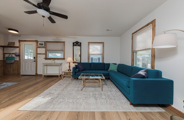 A living room with a sectional sofa, a glass coffee table, a ceiling fan, and a large window with blinds.