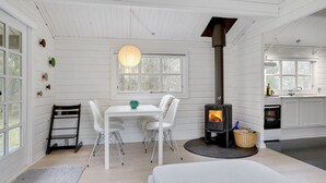 Une pièce aux murs blancs, une table avec des chaises, un luminaire suspendu, une cheminée et un coin cuisine avec des placards et une fenêtre.