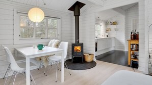 Un intérieur moderne aux tons clairs, avec des éléments en bois peints en blanc, une cheminée centrale, un coin repas comprenant une table et des chaises, et une cuisine dotée de meubles blancs.