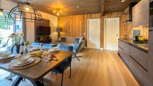 Une cuisine moderne avec des meubles en bois, un coin repas avec une table dressée pour quatre personnes, et un salon avec un canapé et un téléviseur.