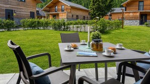 Une cabane en bois avec une table à manger installée à l’extérieur, entourée d’une pelouse bien entretenue et avec une montagne boisée en toile de fond.