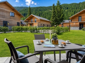 Una cabaña de madera con una mesa de comedor al aire libre, rodeada de un césped cuidado y un paisaje montañoso cubierto de bosque.