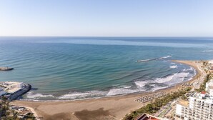 Vista costera con playa, un rompeolas y una zona turística con edificios y palmeras.