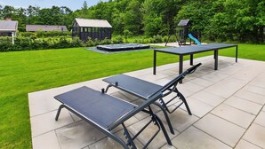 Een terras buiten met een tafel en bankjes, omgeven door een verzorgd gazon en bomen.