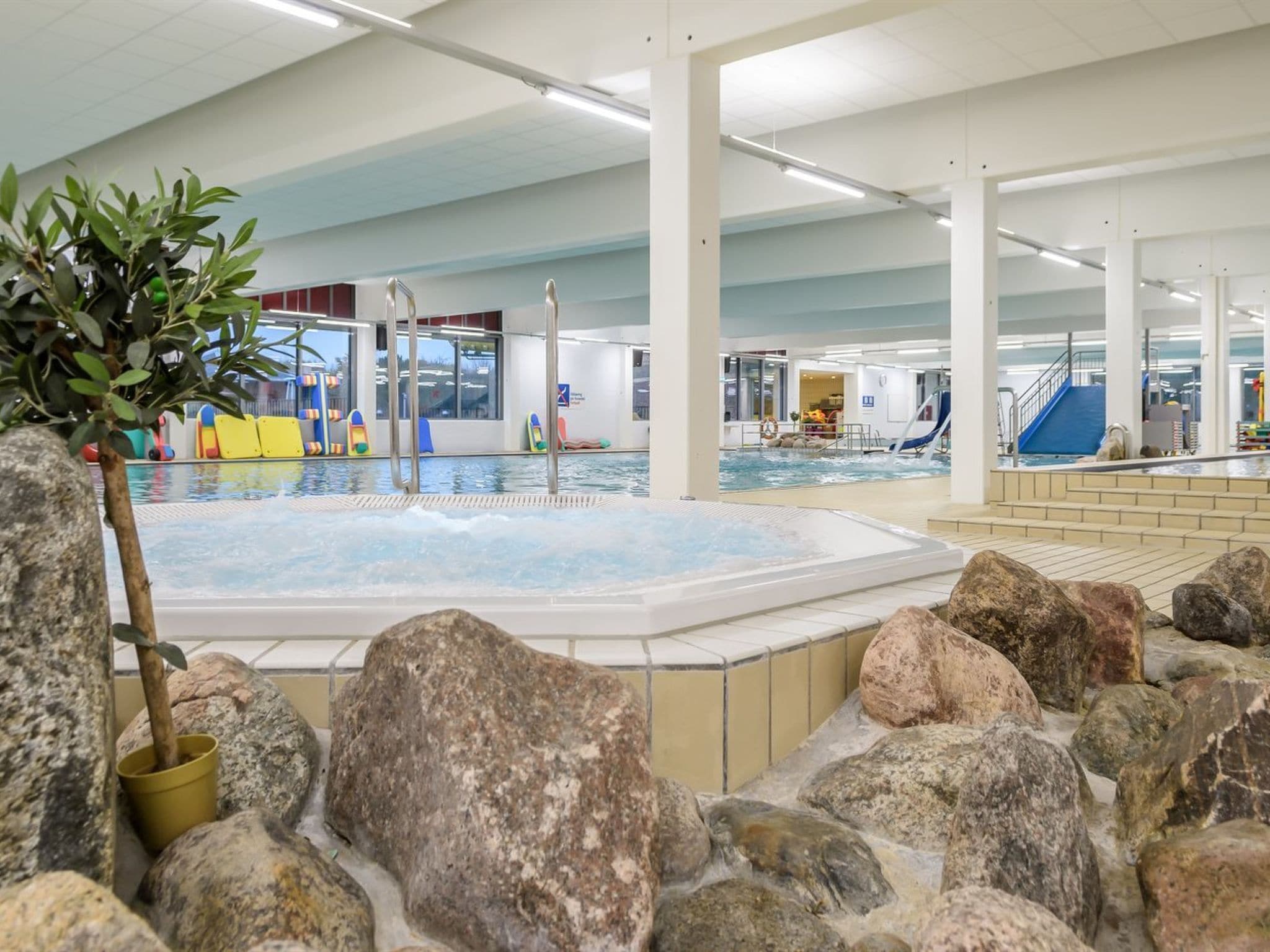 An indoor swimming pool with a hot tub, surrounded by rocks and a potted plant.