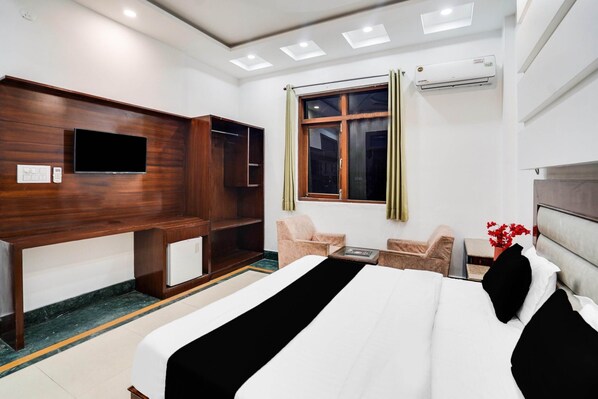 A hotel room with a bed, a TV, a desk, a chair, a small table, and an air conditioning unit.