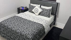 A bedroom with a bed, bedside tables, a nightstand, a window with blinds, and an air conditioning unit.